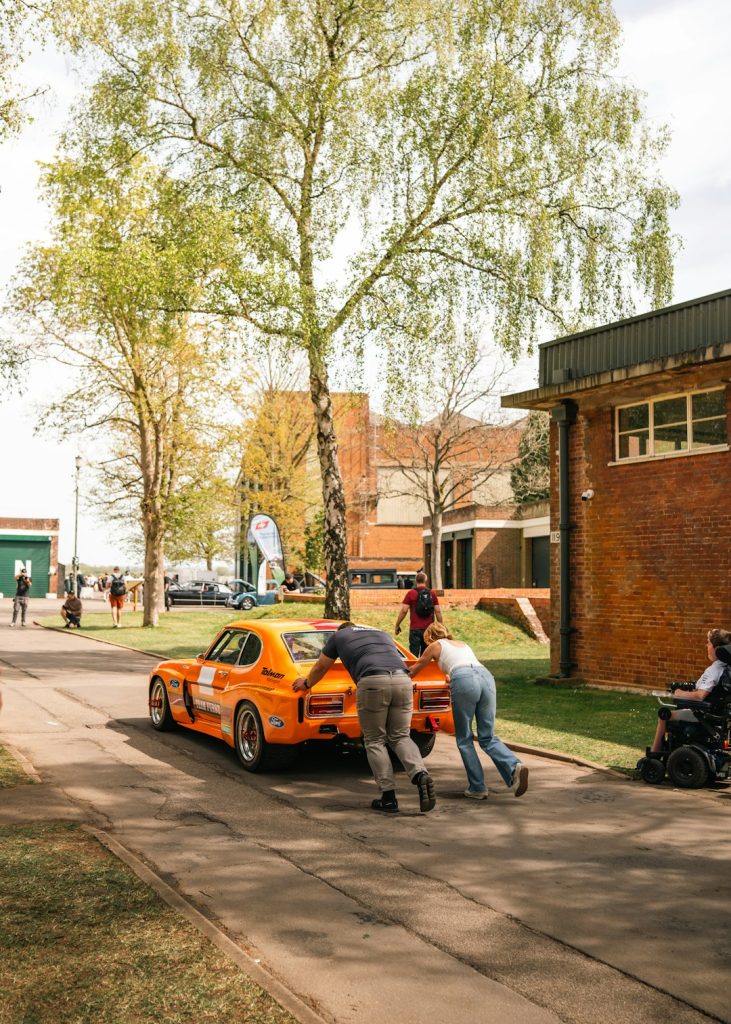 people-pushing-a-bright-orange-vintage-race-car-uhi8q5credy
